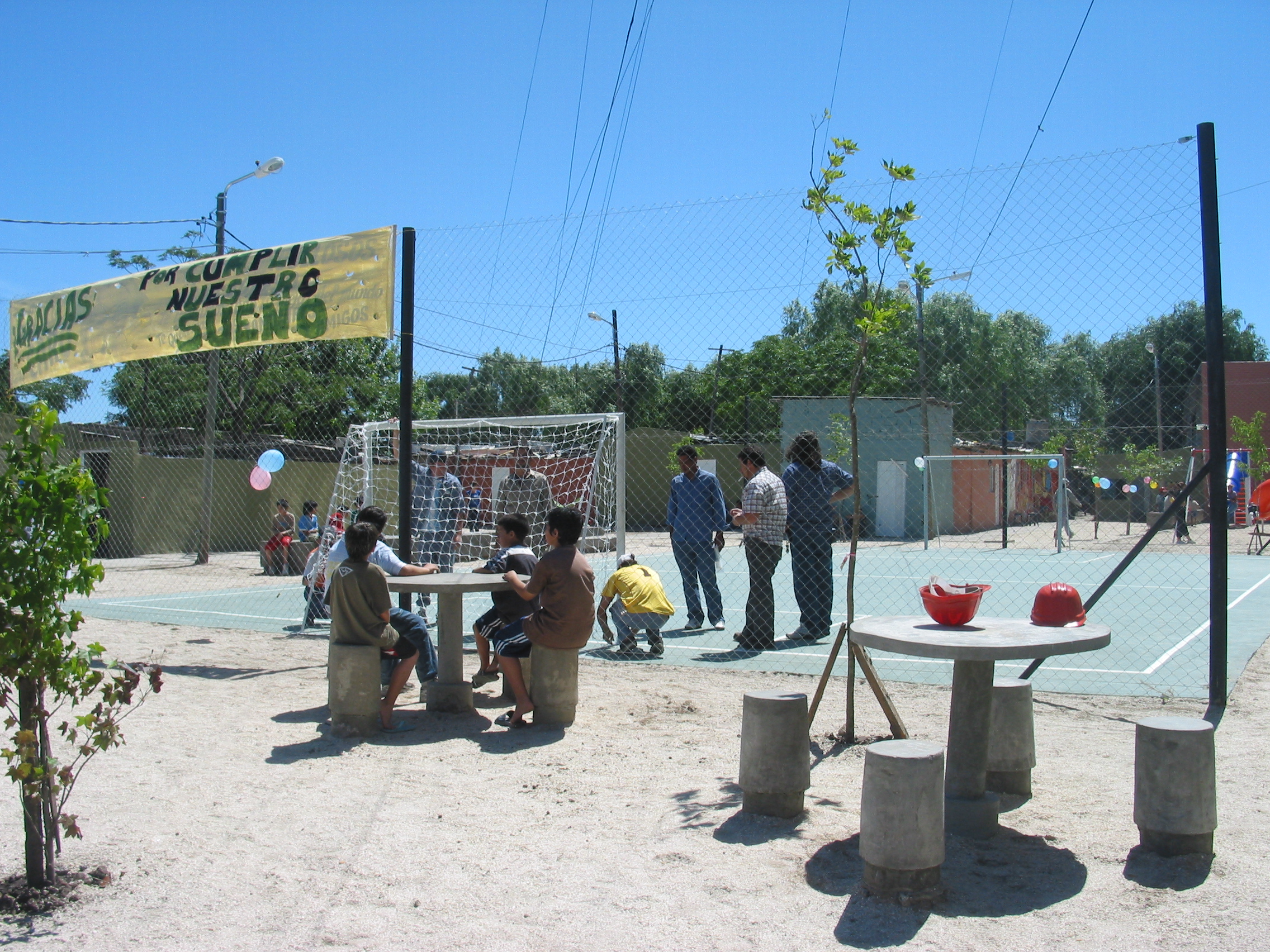 A group of children sitting at a table outside and men repairing a soccer goal at a field.